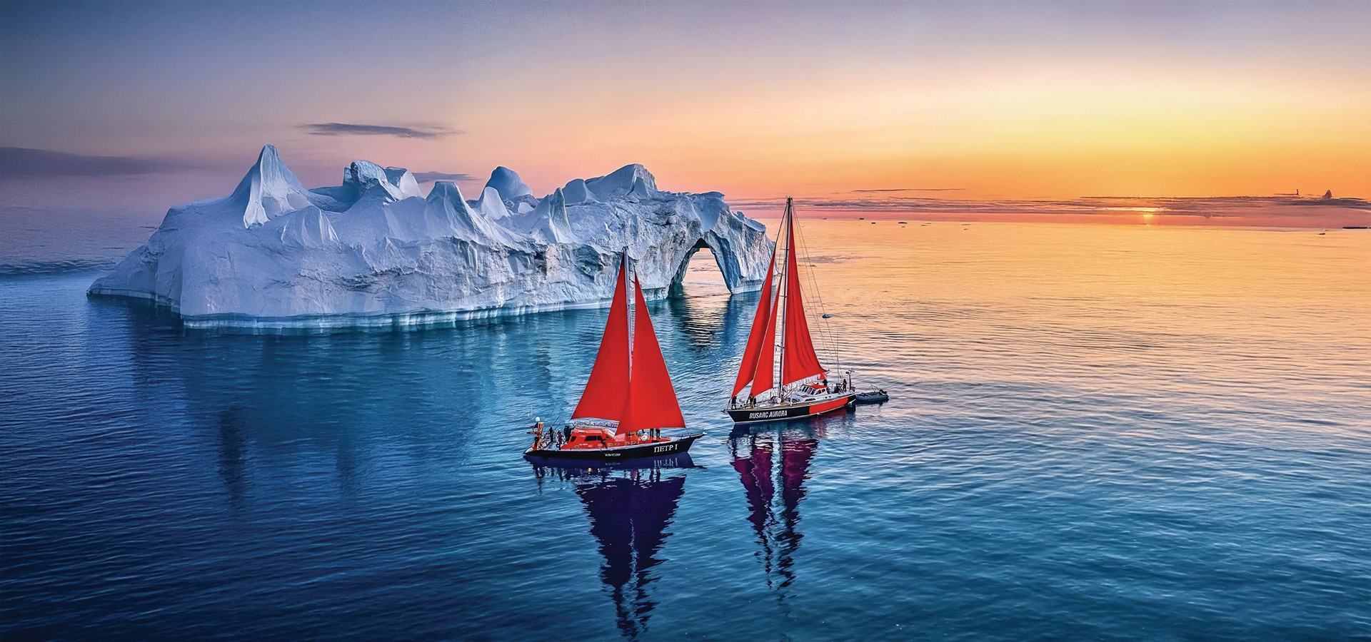 Two sailboats with red sails gliding past an iceberg at sunset